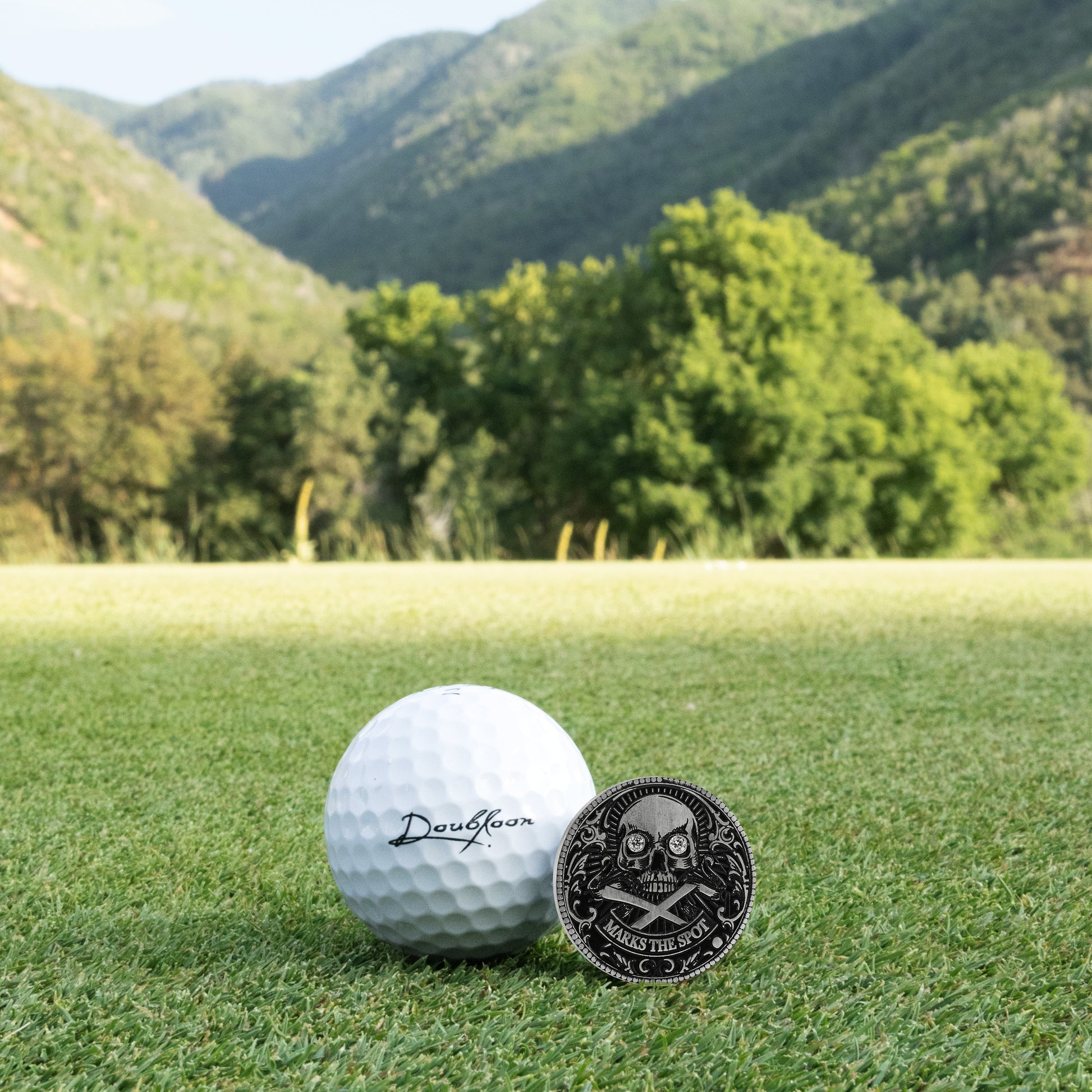 Golf ball and custom luxury golf ball marker with diamonds set in sterling silver and skull design on grass with mountains in the background
